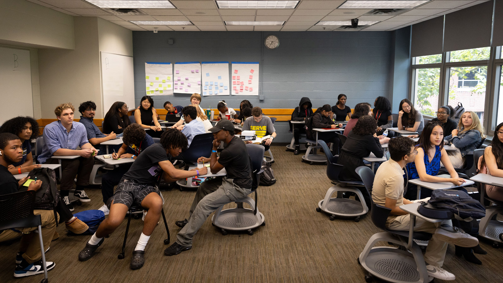 Groups of students work together at circular desks in a well-lit classroom. Some are writing or using laptops, while colorful posters with sticky notes hang at the back of the room.
