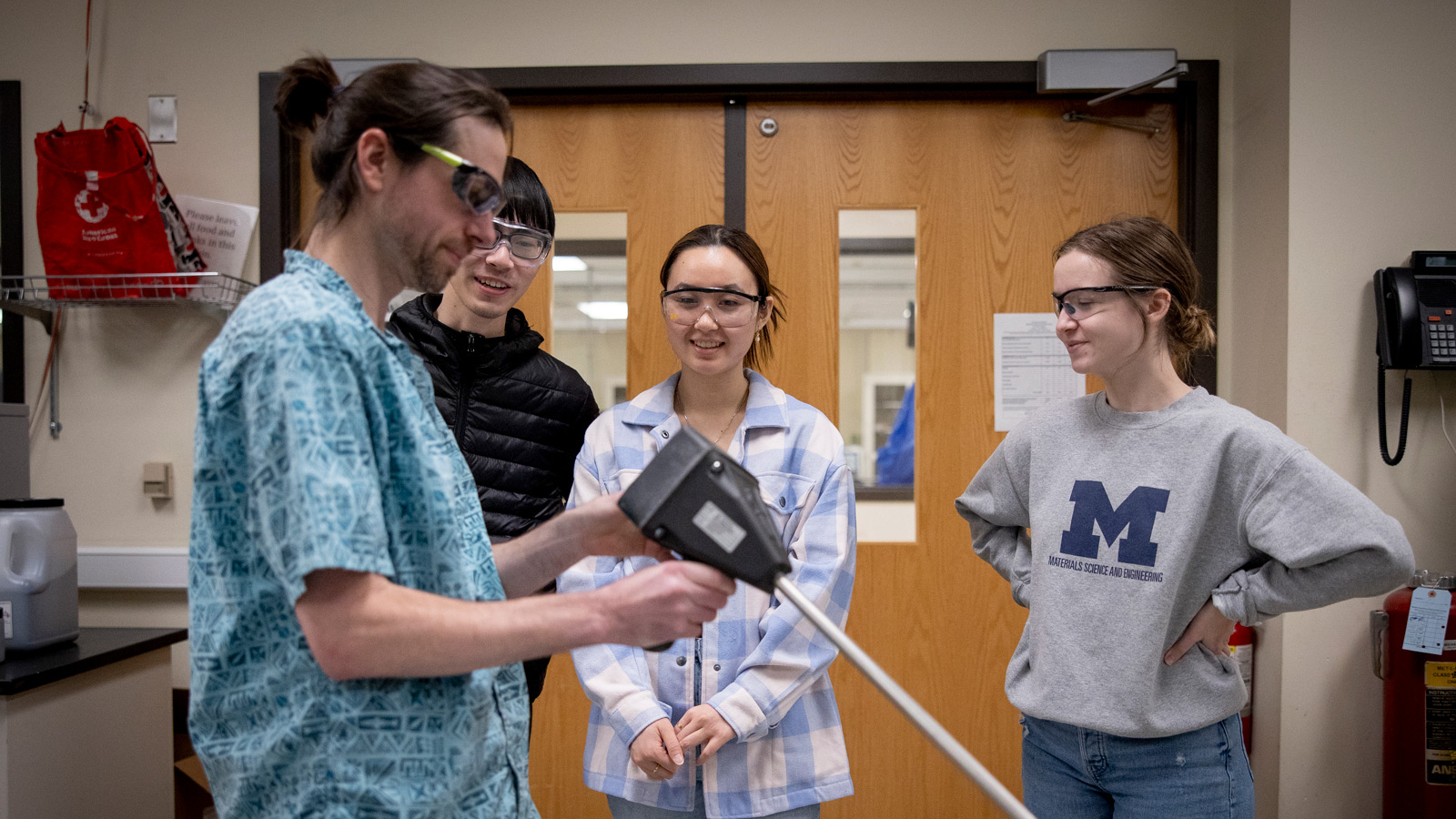 An instructor uses a tool while three students observe in a laboratory, with one student in a Materials Science and Engineering sweatshirt.