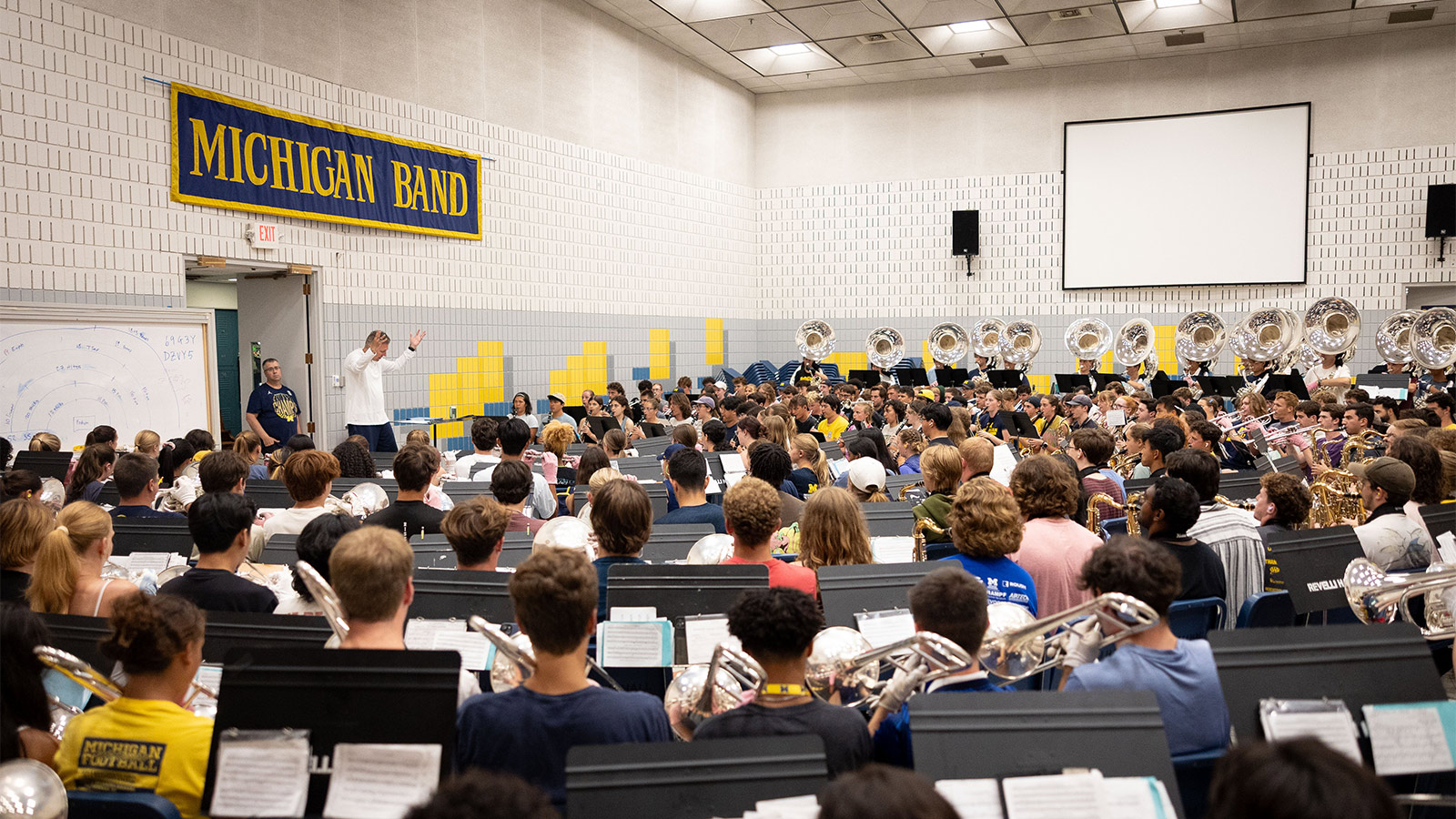 A panoramic view of the Michigan Marching Band practicing in Revelli Hall. A banner that reads MICHIGAN BAND hangs on the wall behind the conductor.