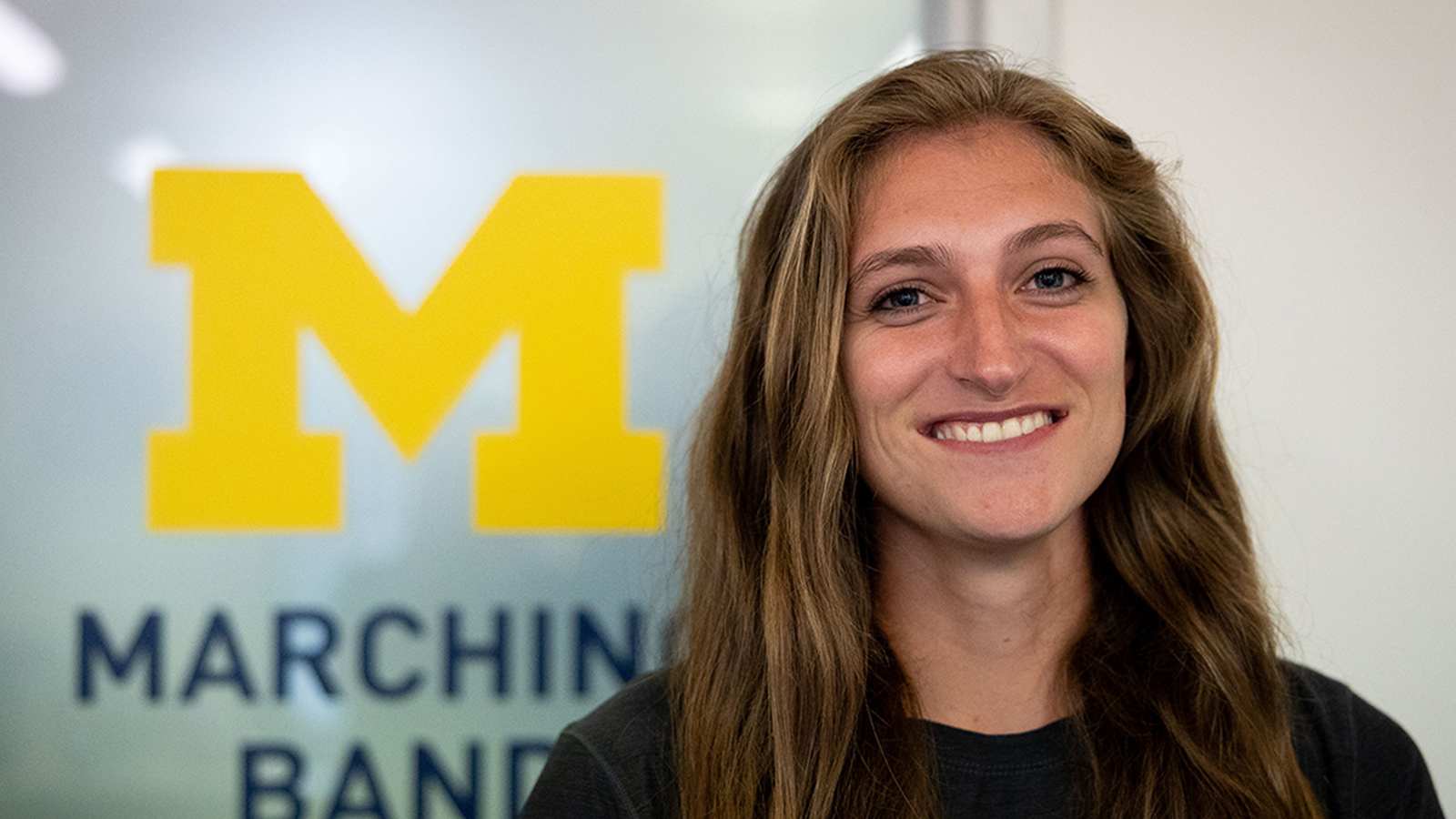 Lauren Streng smiling at the camera in front of a window with the Michigan Marching Band logo in the background.