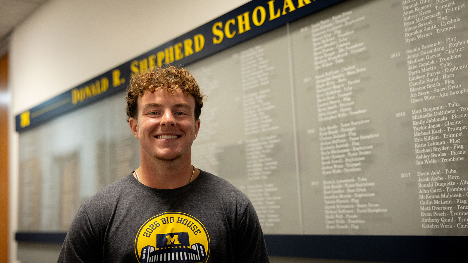Zach McCain standing in front of a wall plaque listing recipients of the Donald R. Shepherd Scholarship.