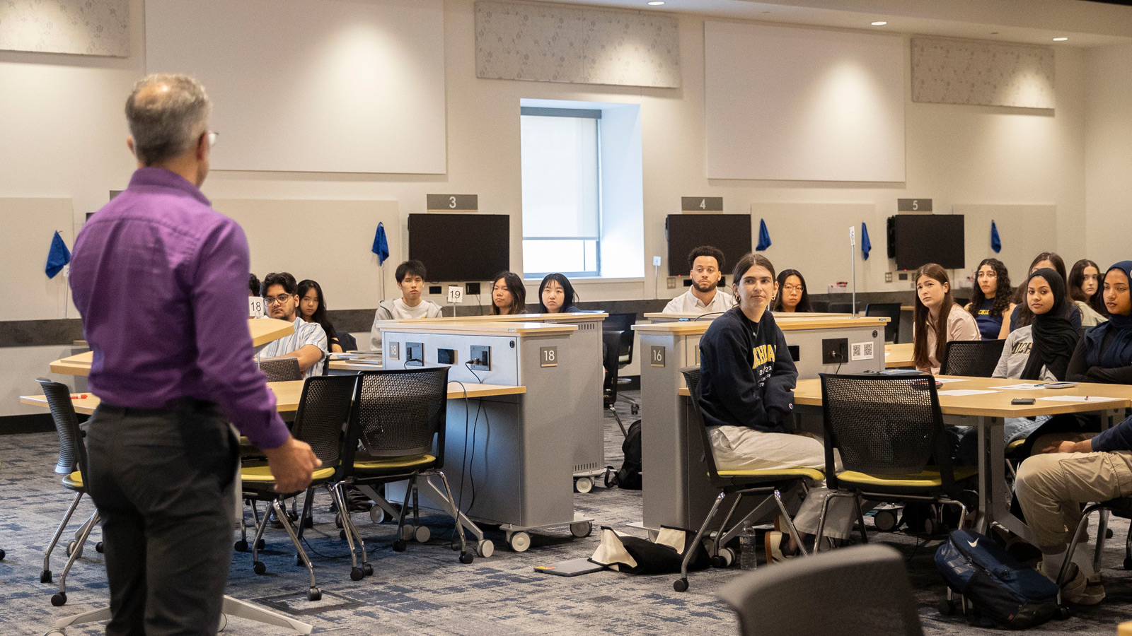 An instructor in a purple shirt addresses a group of students wearing Michigan apparel and casual clothes, seated in clusters at tables equipped with monitors in a bright, high-tech classroom.