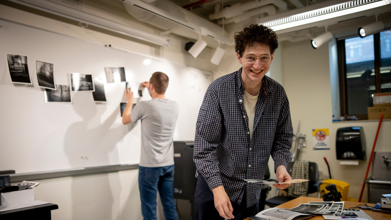 A person in a checked shirt sorts black-and-white photographs at a table while another person attaches similar prints to a whiteboard in a studio setting with exposed pipes and bright lighting.