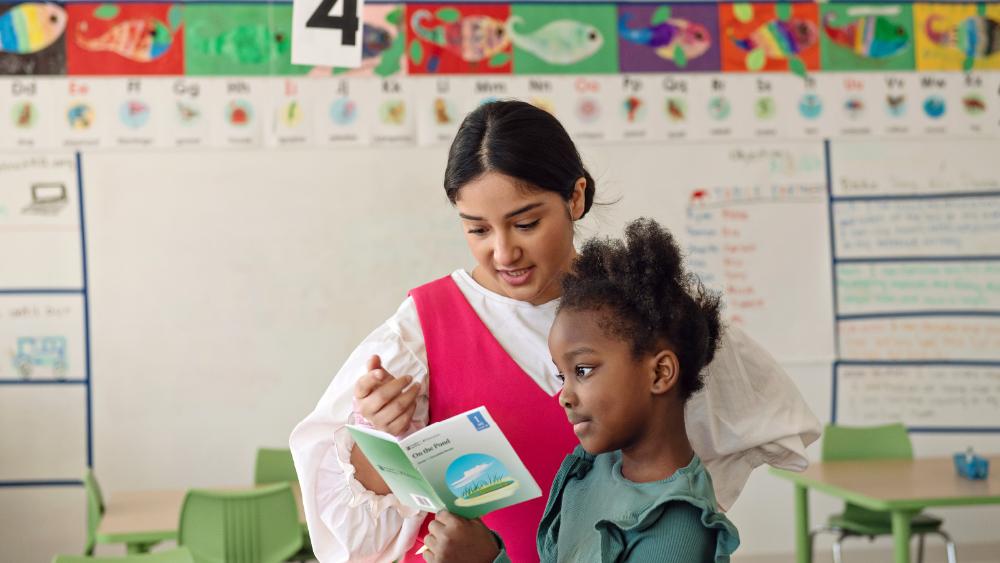 An elementary school teacher helping a young girl to read a book.