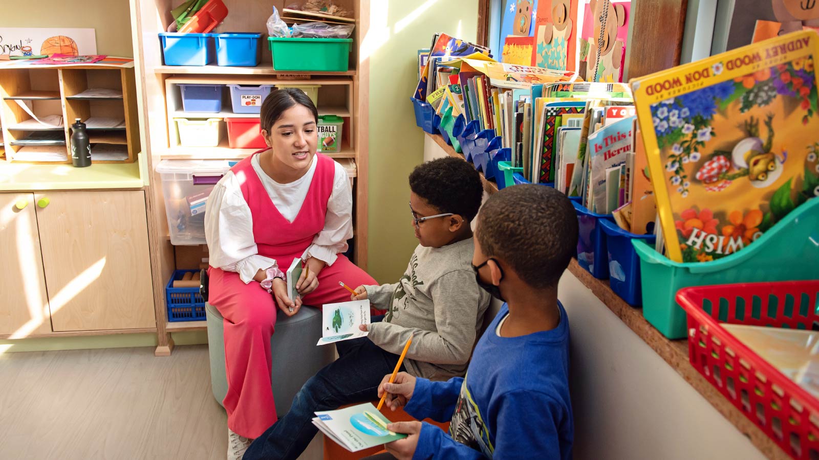 A teacher sitting next to two boys holding books and pencils.