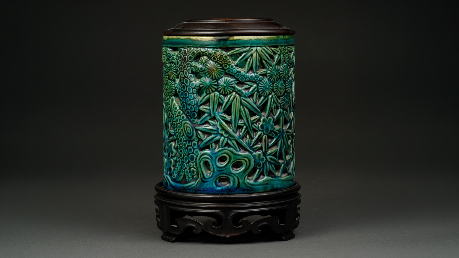 A carved green-and-blue cylindrical vessel sits on a dark wooden stand against a black background.