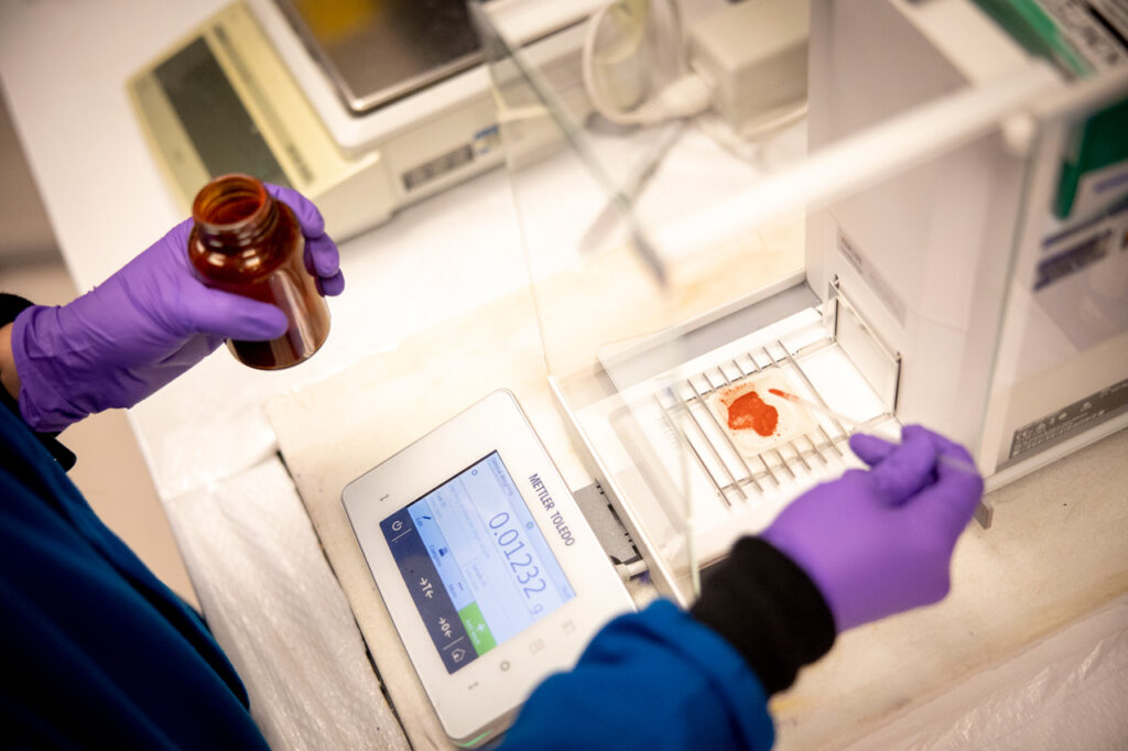 Two hands with rubber gloves on holding a jar and brush above a scale with a liquid sample on it.