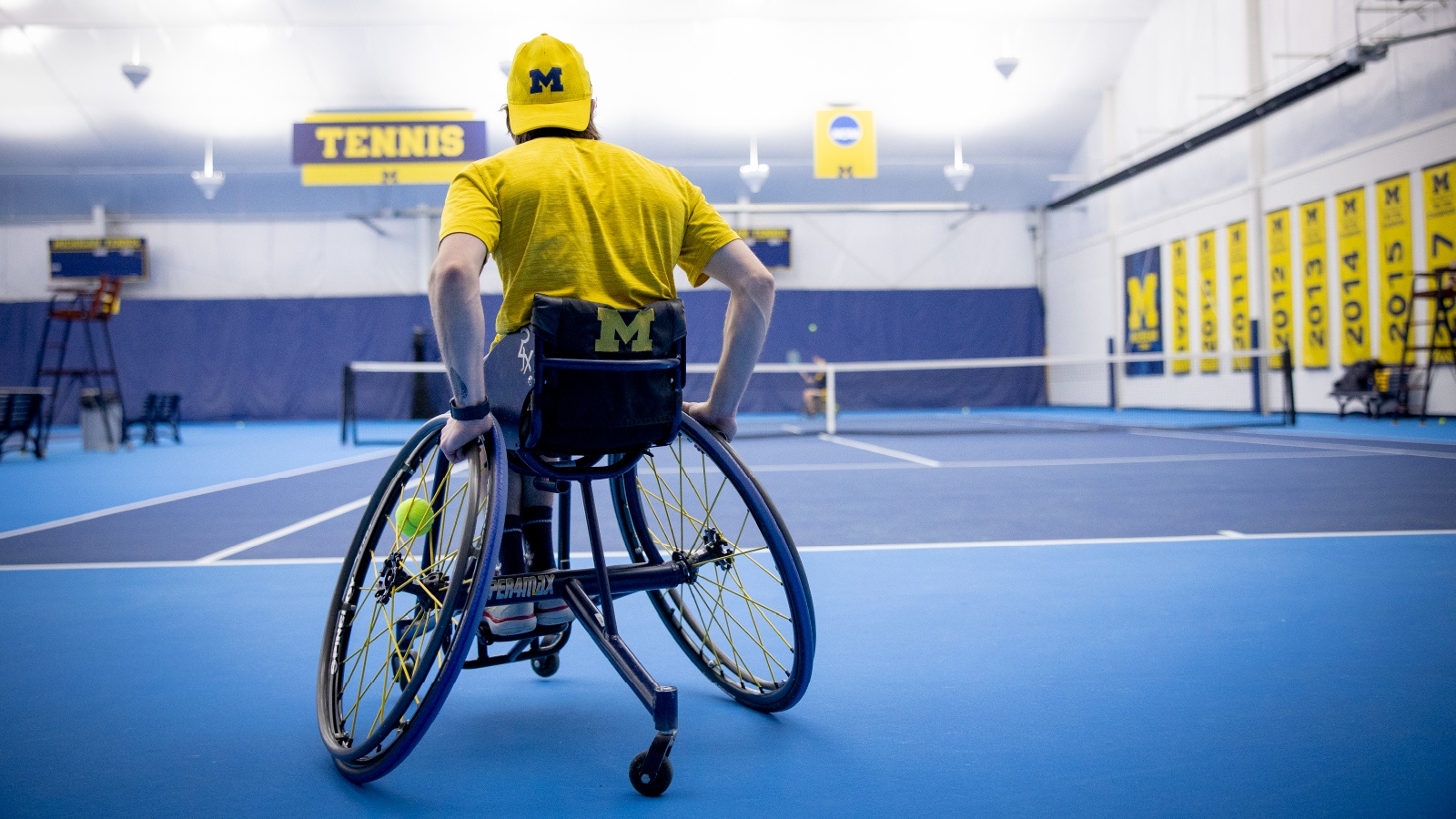 A man in a wheelchair seen from behind with a tennis ball in the spokes on a U‑M indoor tennis court.