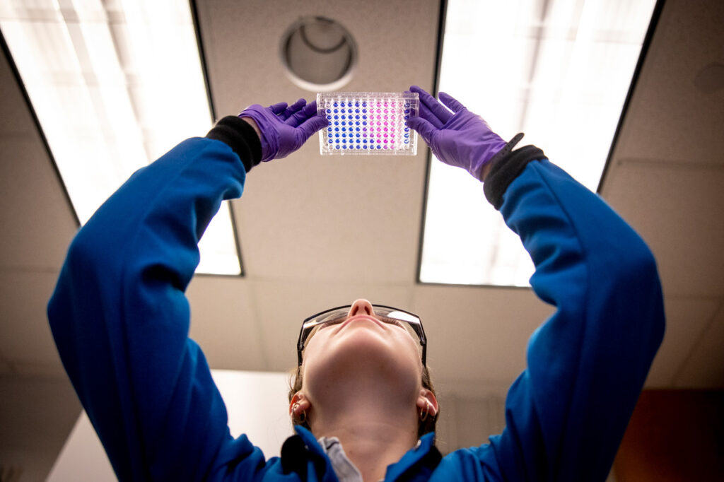 A woman holding up lab test samples under the lights in the room and looking up at them.