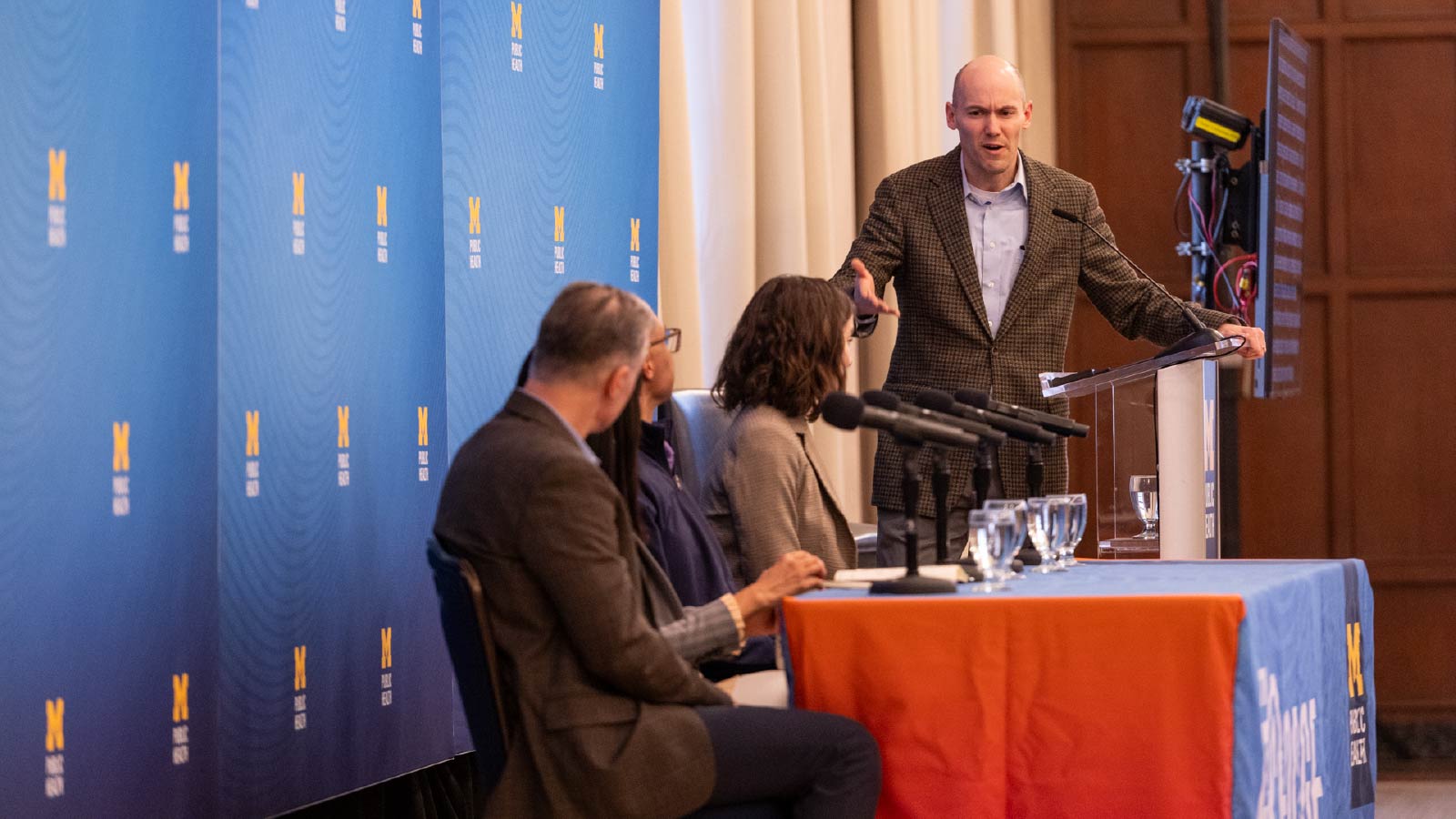 Justin Heinze standing at a podium and moderating a panel. He’s facing toward four panelists who are sitting at a table. 