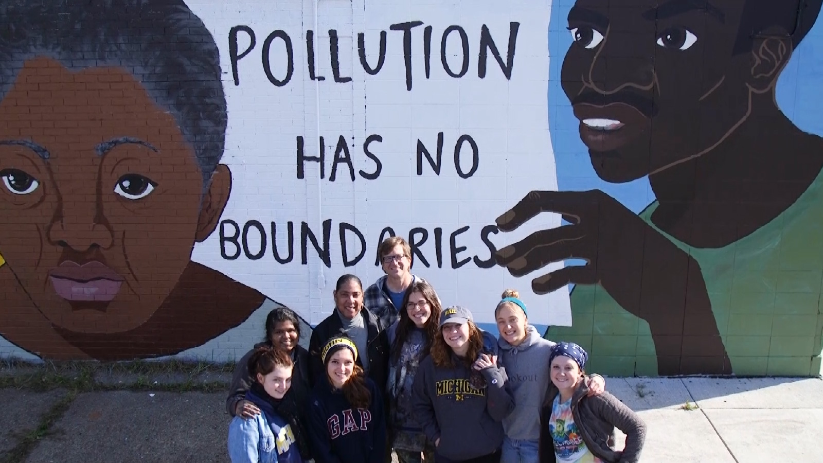 A diverse group of people stands in front of a mural that features two people and the words “Pollution has no boundaries.”