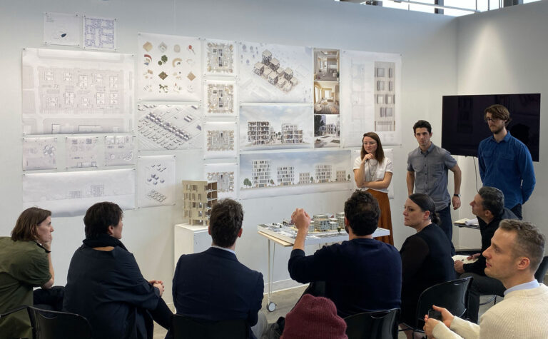 A classroom with drawings on a wall, students standing, and reviewers seated discussing their work.