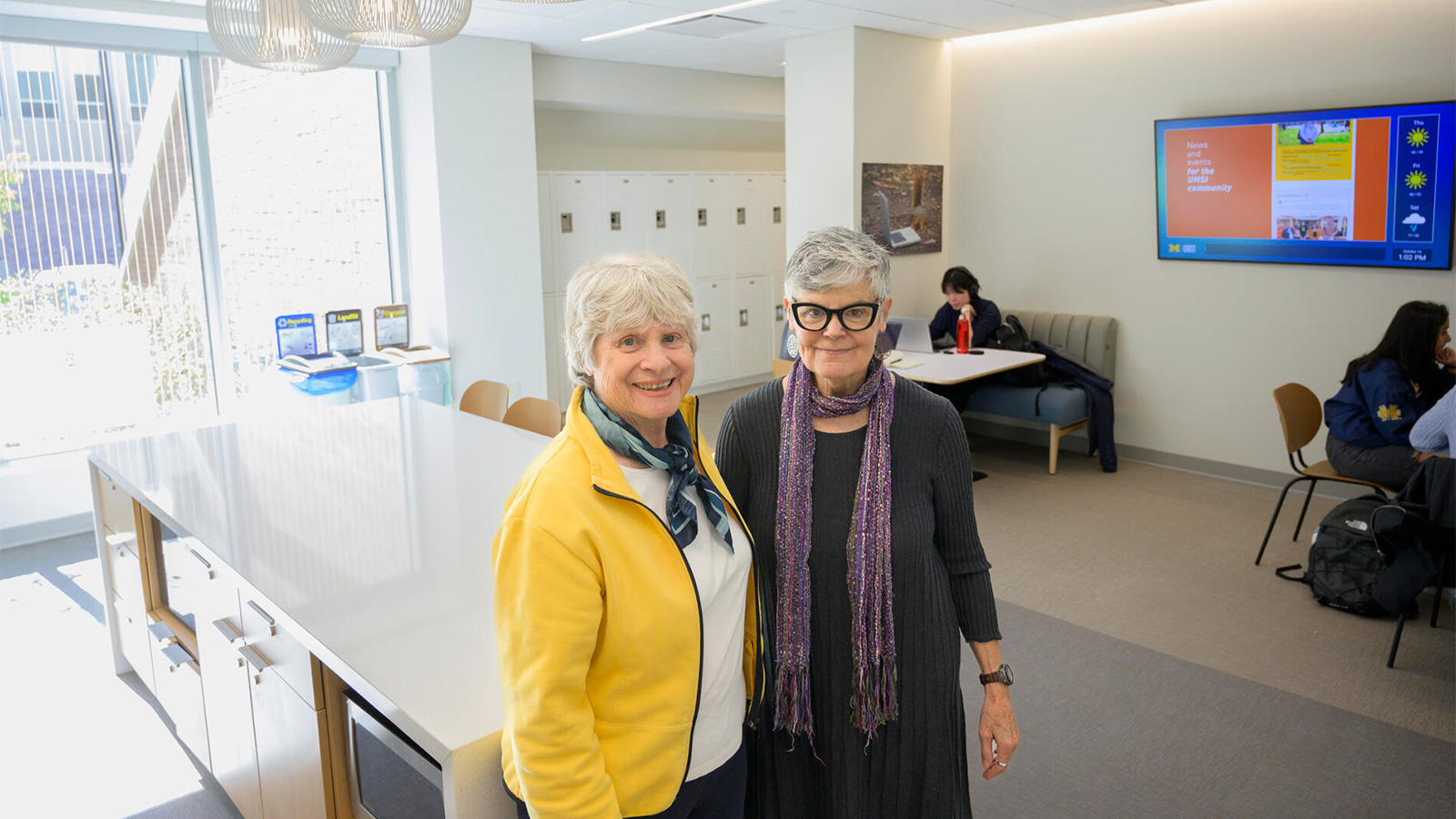 Quackenbush and Kenlee Ray posing in the student lounge, with a wide kitchen island and hanging light fixtures behind them. Students seated at tables and a row of lockers are visible in the background.