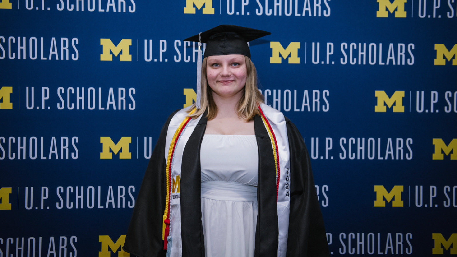 Molly Helminen wearing a cap and gown in front of a background that reads ‘M’ U.P. Scholars 