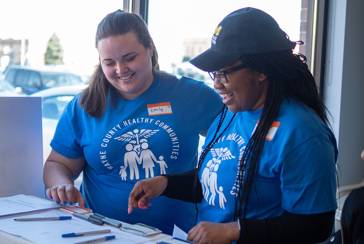 A photo of two people wearing shirts that say “Wayne County Healthy Communities.” One is wearing a U‑M hat.