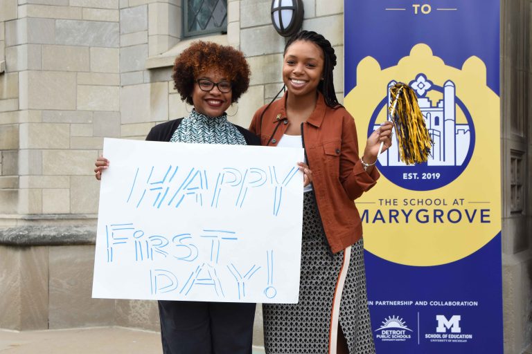 Two doctoral students in front of the School at Marygrove holding a sign that says "Happy first day!"