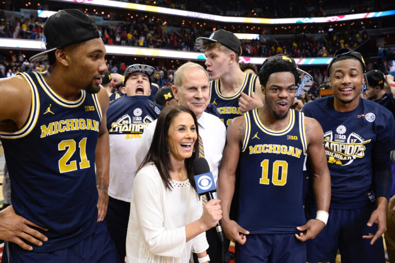 Wolfson is holding a microphone and standing next to former Michigan coach John Beilein and members of the men's basketball team.