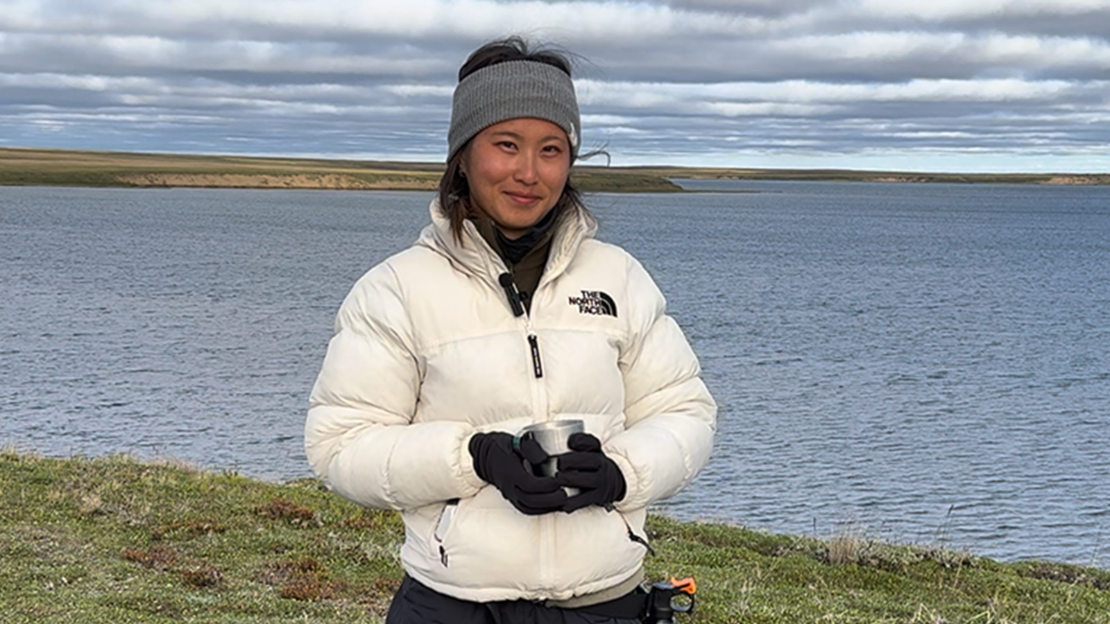 A woman standing outside, in front of a large body of water. She’s wearing a white coat and a gray winter headband. 