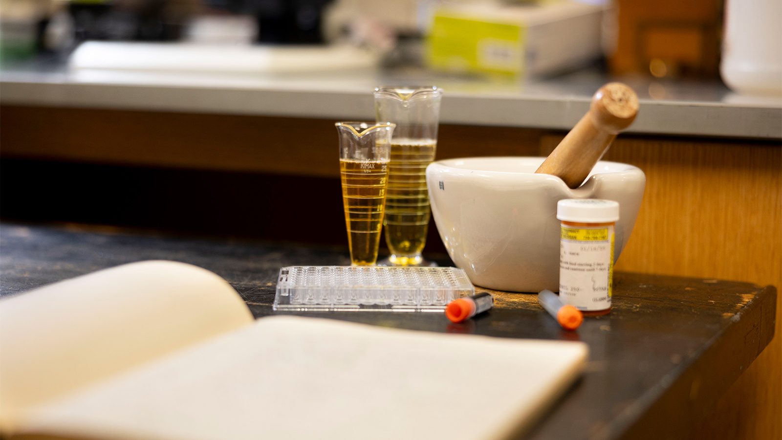 A laboratory workbench holds two graduated cylinders with yellow liquid, a white mortar and pestle, a vial, a test tube tray, a small orange-capped tube, and an open notebook in the foreground.