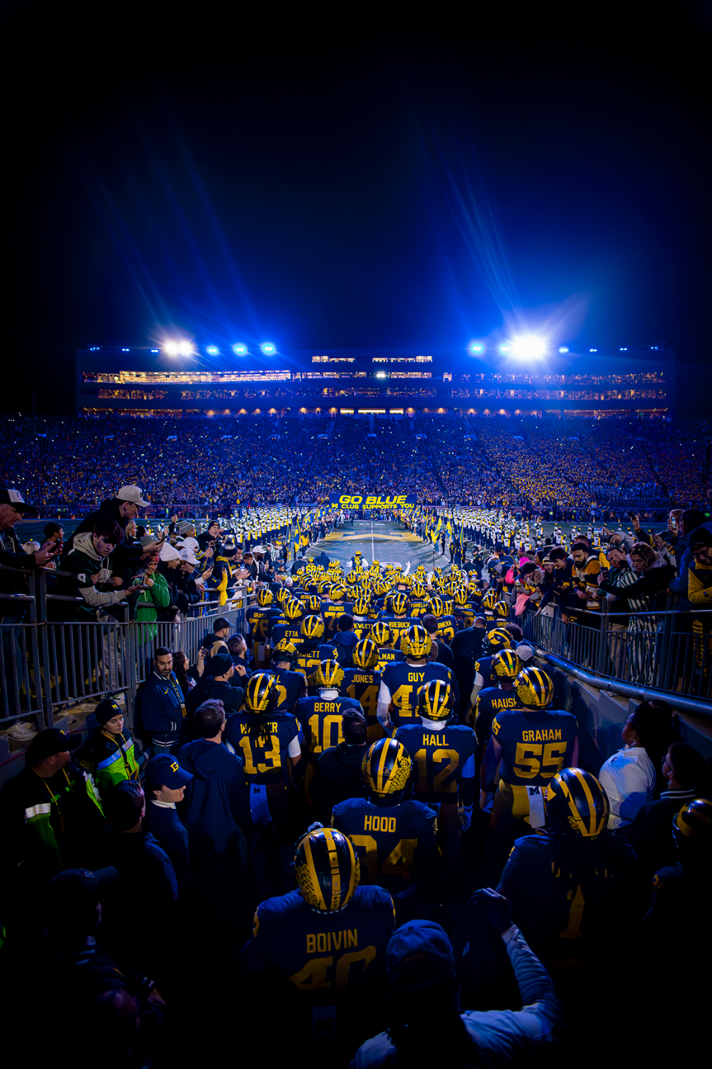 Michigan football players running out of the tunnel onto the field at the Big House, surrounded on both sides by bystanders in the stands.