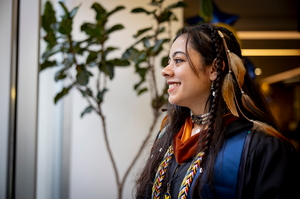 A student wearing a cap and gown looks out of a window smiling.