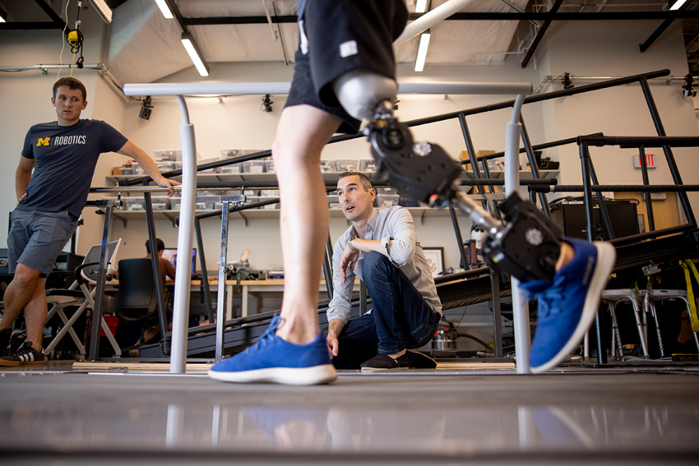 In focus, a person with a prosthetic robotic leg walks across the floor. In between that person's legs, in the background, a second person is kneeling on the floor. To the left, a third person is standing, watching the demonstration.