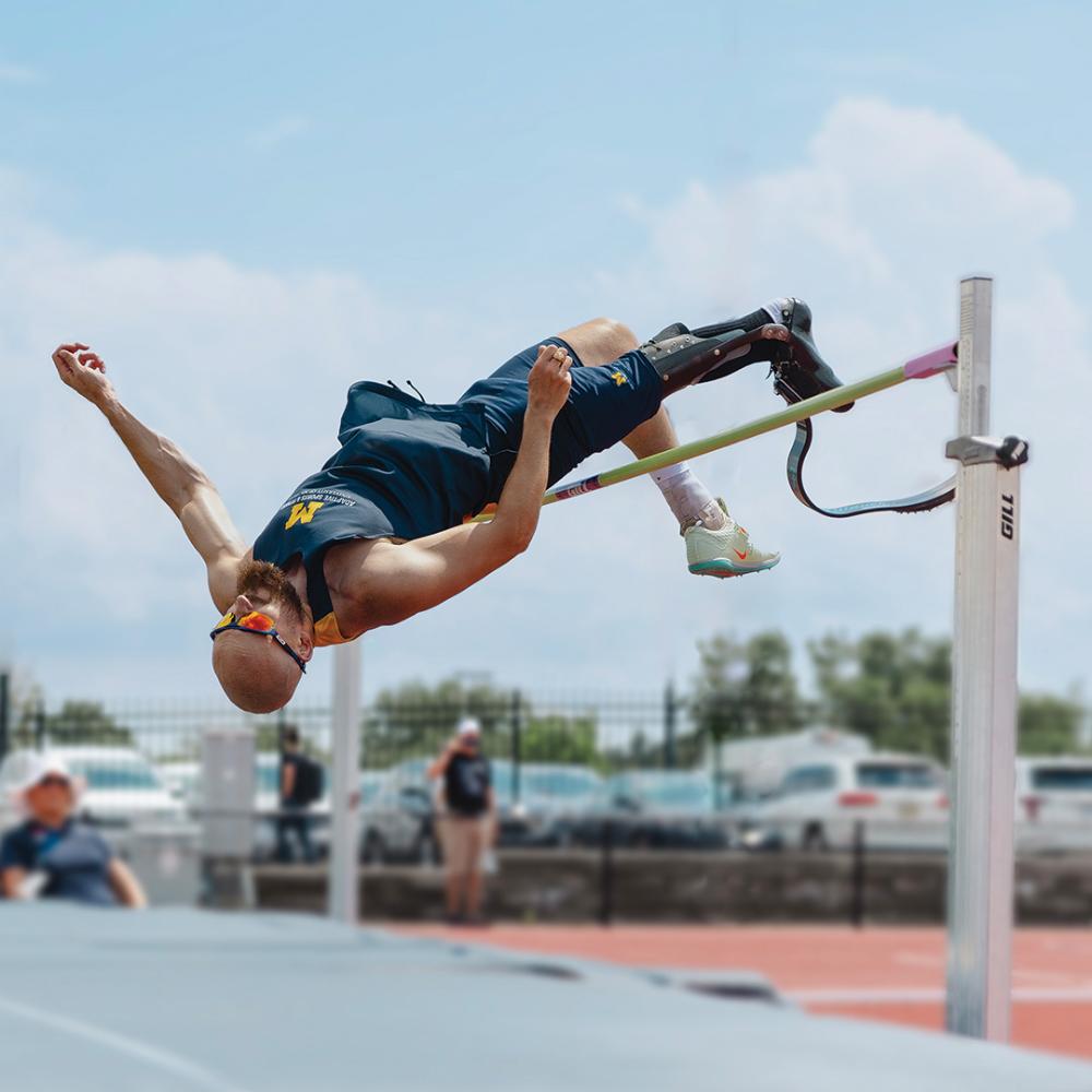 A male high jump athlete in a U-M jersey with a prosthetic leg jumping over a horizontal bar.