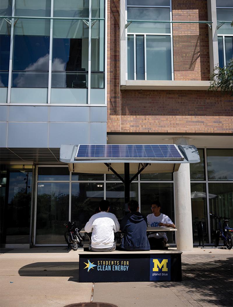 A few people are sitting at a table with a solar panel above it. A bench that two of them are sitting on reads “Students For Clean Energy” and also features a block M with the words “Planet Blue” under it. The table is on a sidewalk in front of a building with a brick, cement, and glass facade.