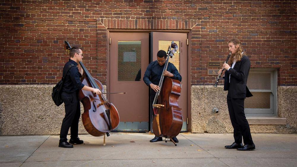 Three young men playing musical instruments, one playing a cello, one playing a double bass and one playing a clarinet, standing in front of doors in the side of a building.