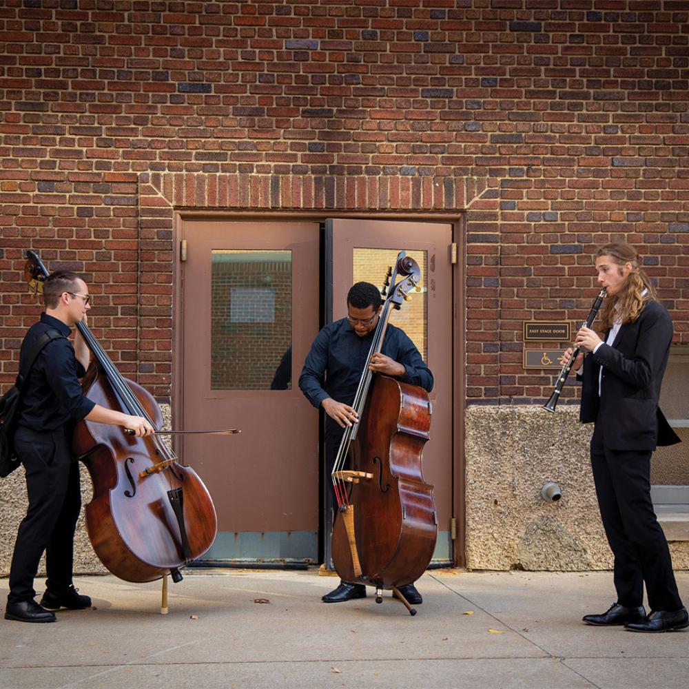 Three young men playing musical instruments, one playing a cello, one playing a double bass and one playing a clarinet, standing in front of doors in the side of a building.