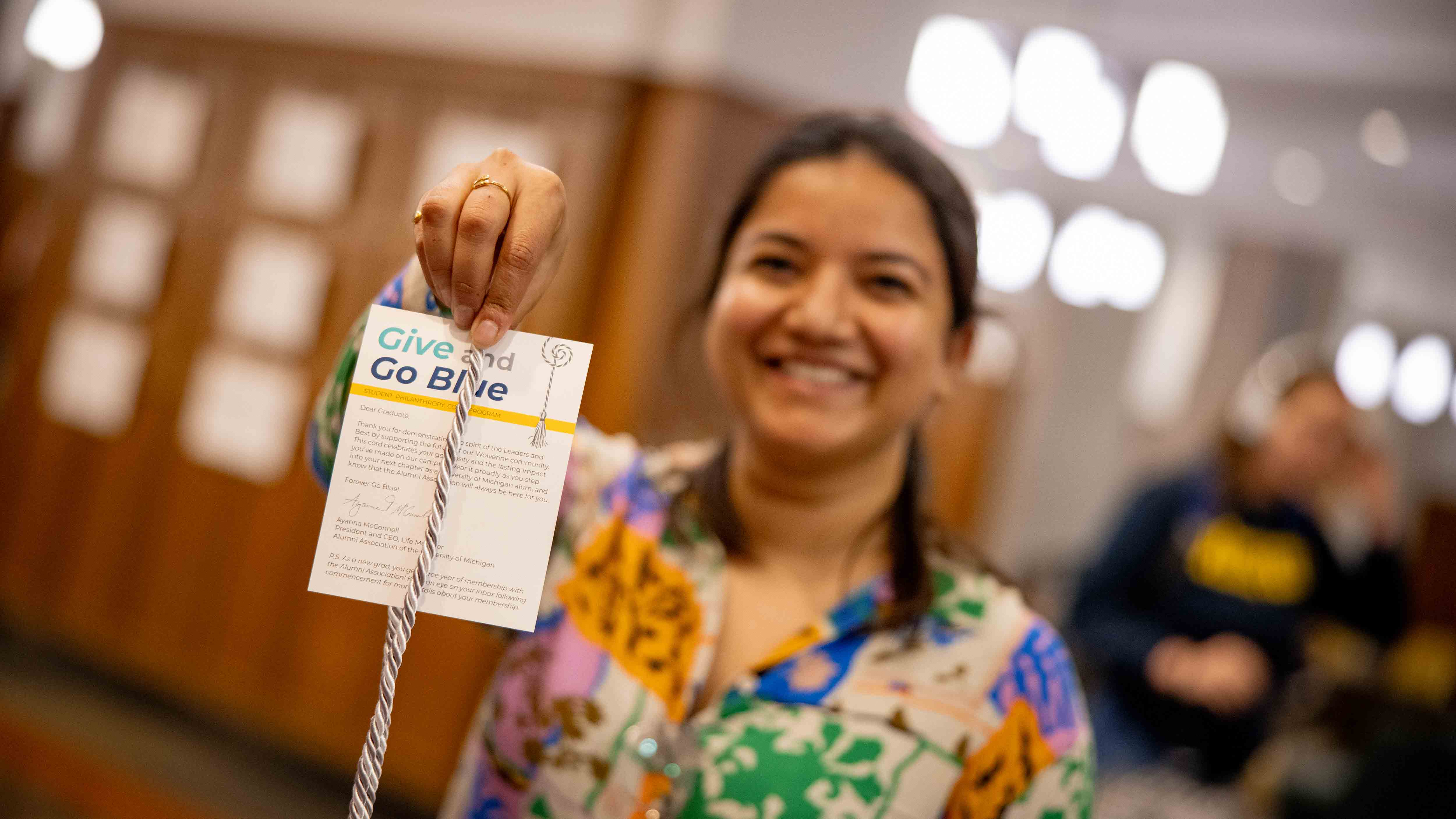 A student out of focus holds up a Student Philanthropy graduation cord to the camera, in focus. Attached to the cord is a thank you note, with large text at the top 
that reads Give and Go Blue.