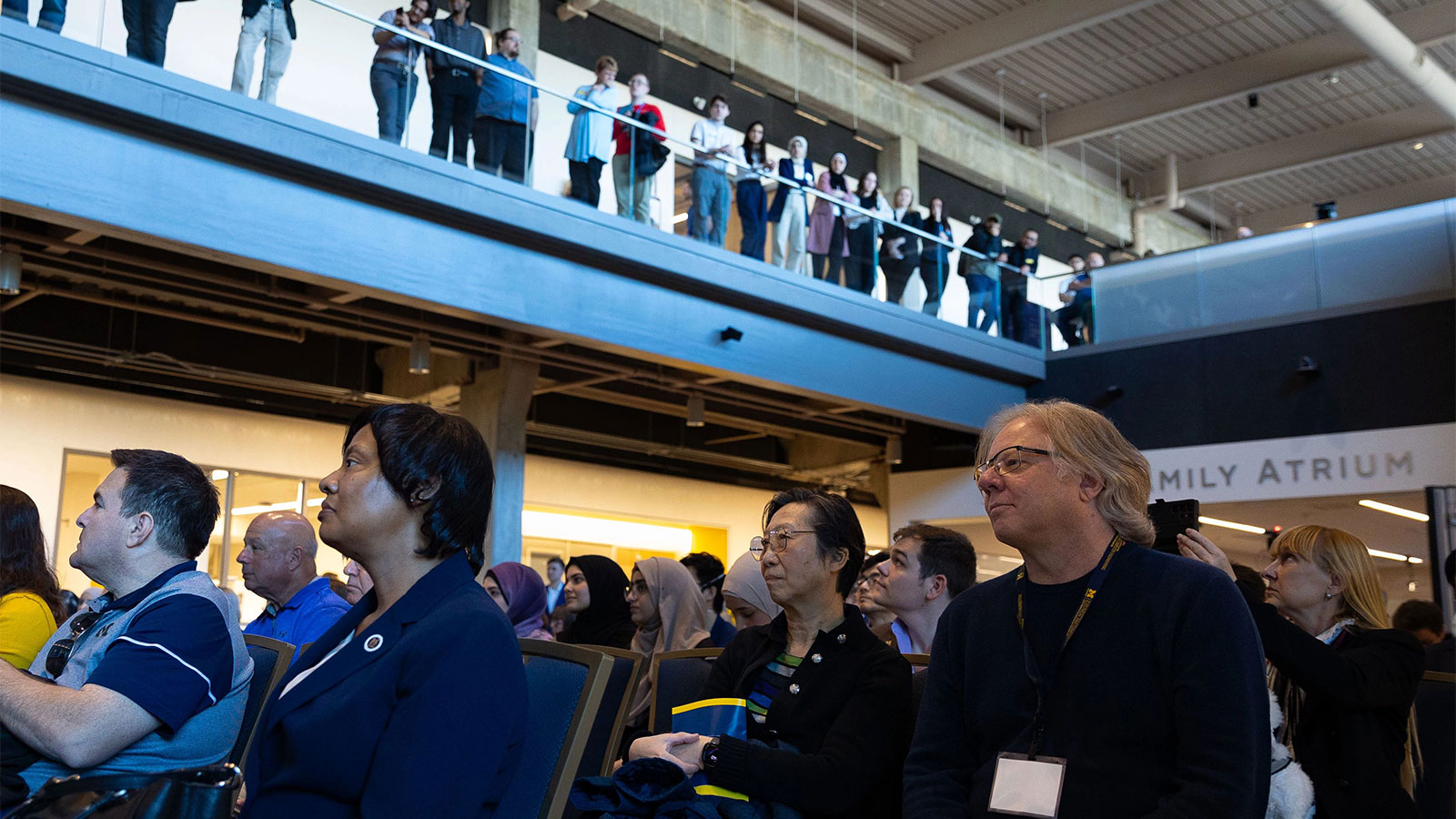 A crowd sitting in a building atrium with other people looking down from a gallery.