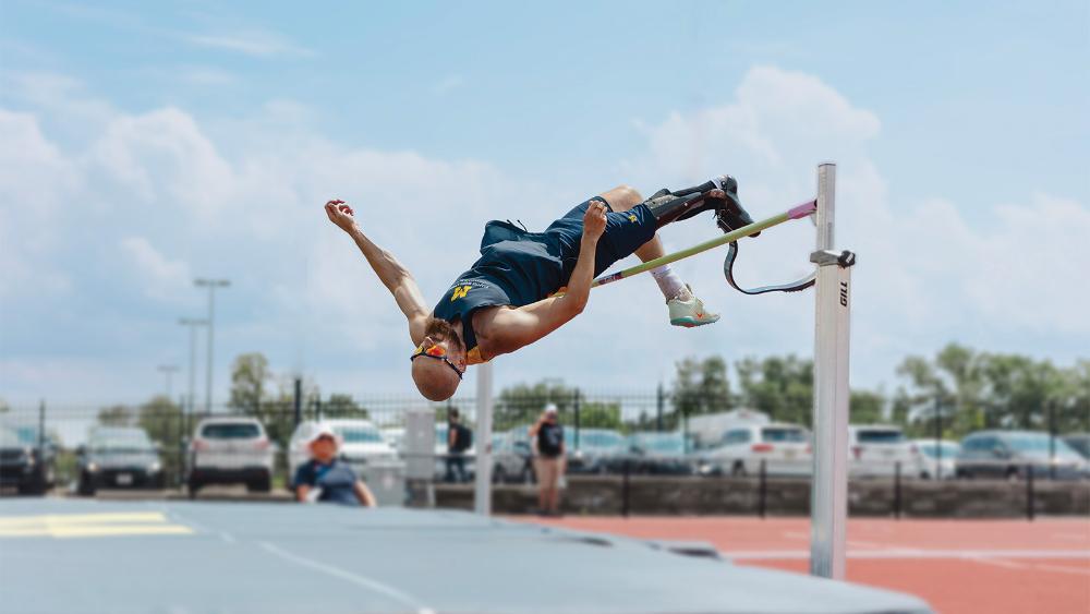 A male high jump athlete in a U-M jersey with a prosthetic leg jumping over a horizontal bar.