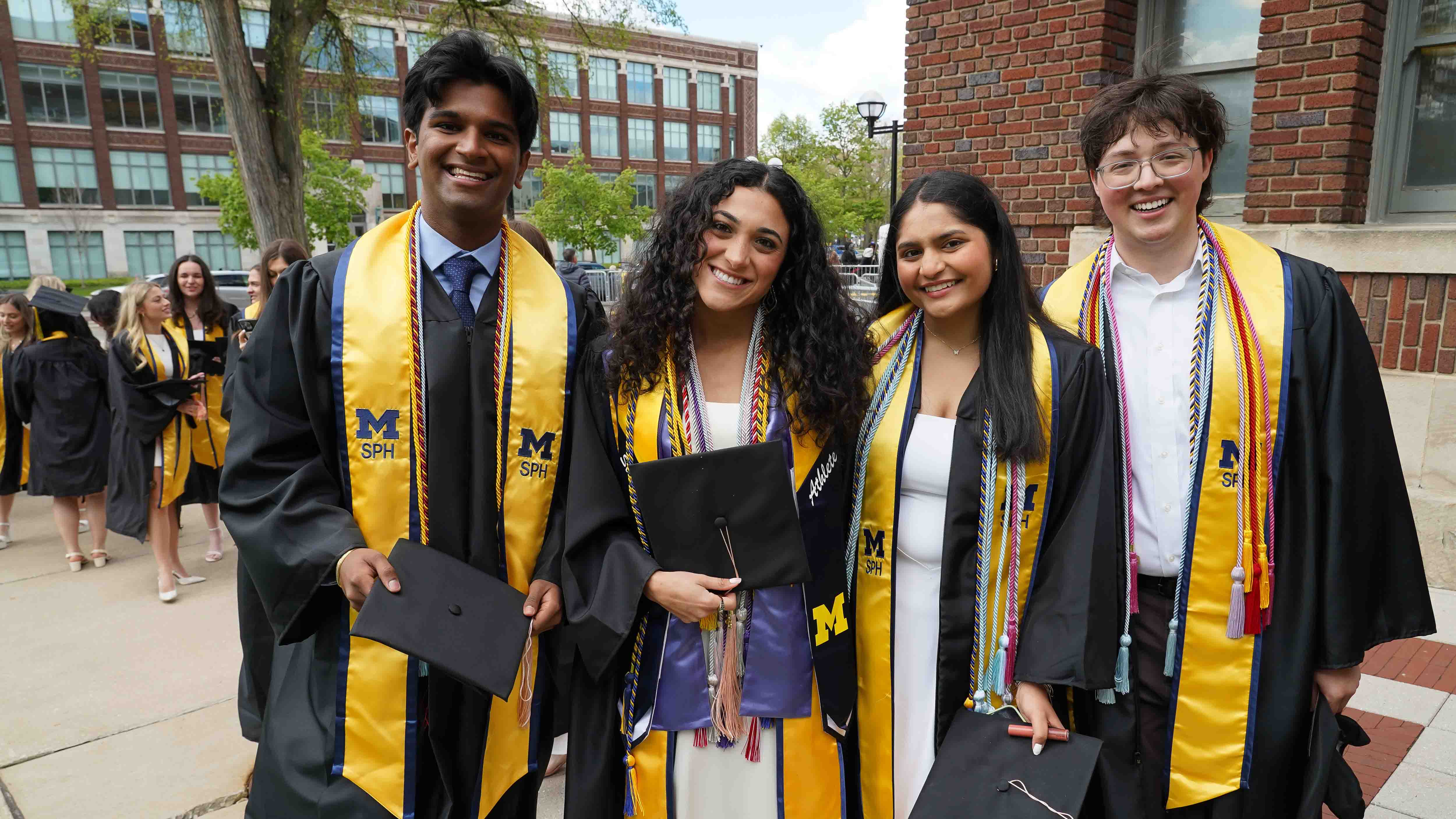 Four School of Public Health graduates stand in a line holding their graduation caps, wearing yellow SPH stoles, and smiling at the camera outside Hill Auditorium in Ann Arbor.