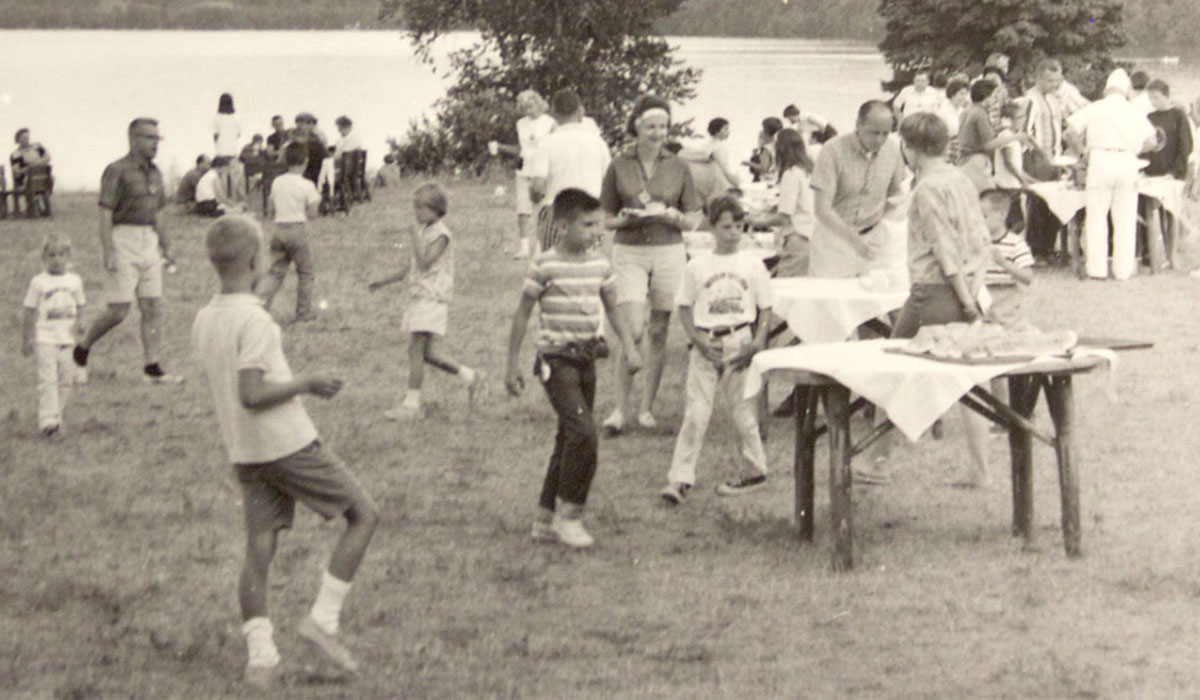 Historic photo from Camp Michigania where people are gathered at a lakeside picnic, with tables of food and children playing on the grass.