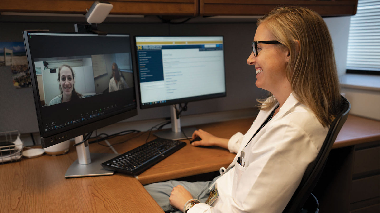  A woman in a lab coat sits at a desk with two computer monitors talking with a patient on Zoom.