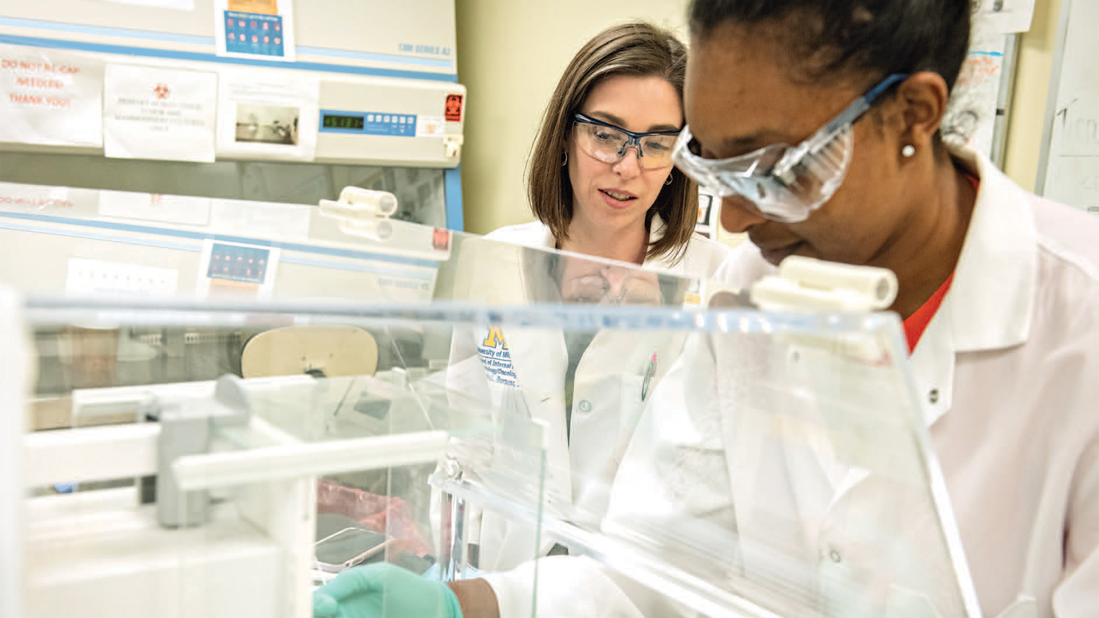 Two women in lab coats and protective eyewear using lab equipment.