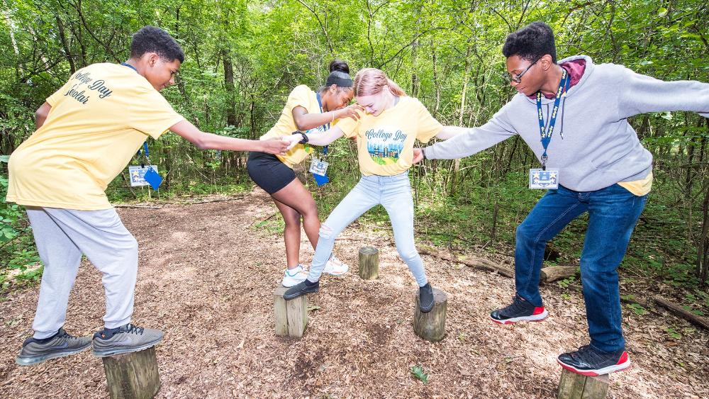 Four students balance on logs in a woodland clearing