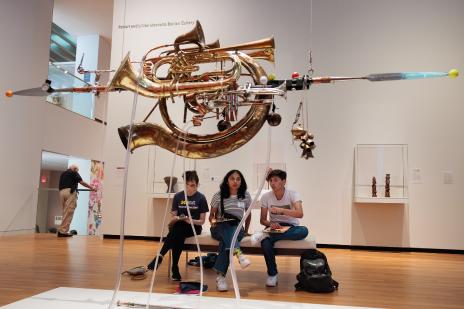 Three students with clipboards and pens sit on a bench, all looking at a hanging sculpture in one of the UMMA galleries