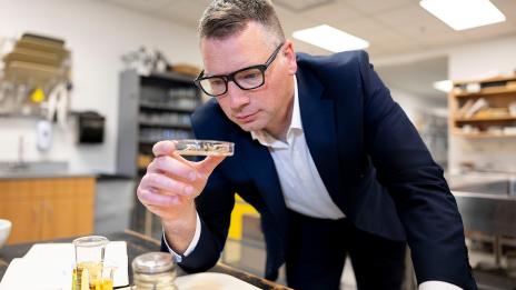 Timothy Cernak in a dark blue suit leans forward, closely inspecting a petri dish while standing in a laboratory with shelves in the background.
