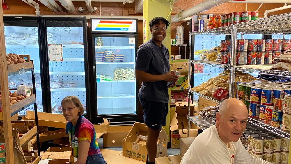 Three people are organizing and stocking shelves in a food pantry. One person is kneeling in front of metal shelves filled with canned goods and pantry staples, another is crouched down sorting sweet potatoes from a cardboard box, and the third is standing near refrigerators in the background. Open boxes and bags of food are scattered around, and the setting has visible pipes and a low ceiling.