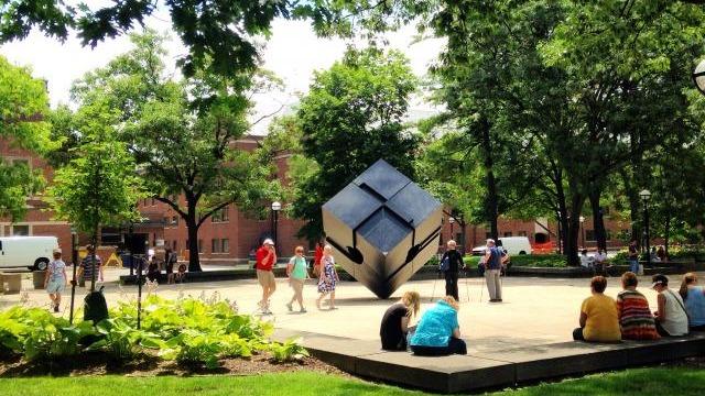 A large, black rotating cube sculpture sits in the center of a paved plaza surrounded by green trees and campus buildings. Several people are gathered around the sculpture, walking nearby, or sitting on low concrete walls bordering the plaza. The scene is bright and sunny, with lush landscaping and a lively campus atmosphere