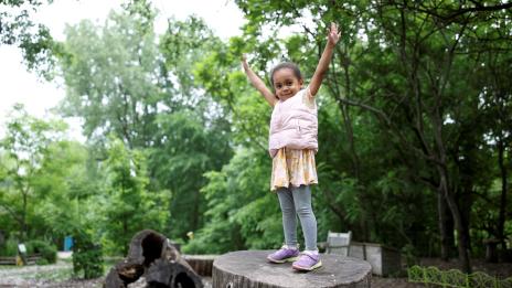A young girls stands atop a tree trunk, surrounded by tall, green trees, at the Gaffield Children's Garden with both arms raised above her head. She is having fun.
