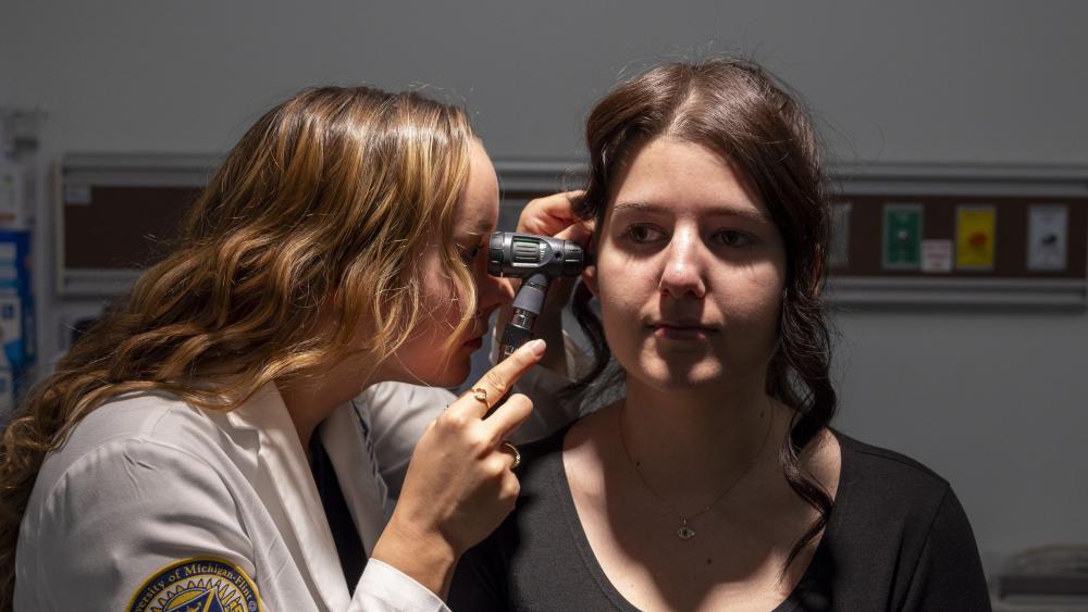 A PA student wearing a white coat uses an otoscope to check the ear of a patient. 