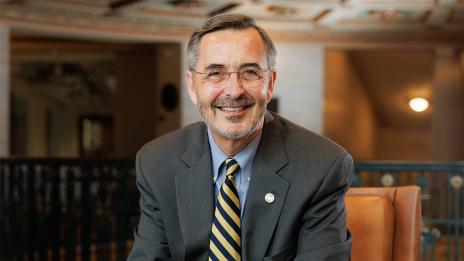 Grasso wearing a gray suit and a yellow-striped tie sits in an ornately designed room featuring warm lighting and decorative architecture.