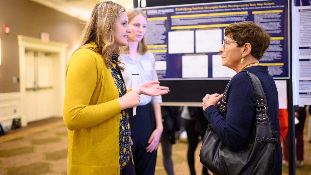  Nursing student explains their research day poster to a guest visitor who listens intently. 