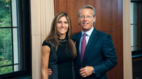 Nate and Catherine Forbes stand side by side, dressed formally, inside a room with wood panels and large windows.