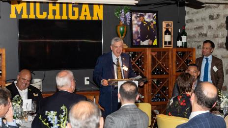Ron Weiser stands at a podium addressing a seated group in a room decorated with Michigan memorabilia.