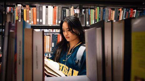 A student in university gear reads a book among library shelves.