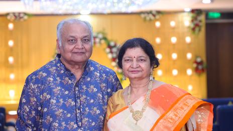 Ashok and Sarita Jain standing together at an event, wearing traditional clothes, with a bright, decorated background.